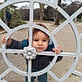 child, curious, decorative_metal, face, hands, hood, jacket, metal_gate, outdoors, park, pavement, portrait, public_park, serious_expression, shoes, standing, toddler, trees, winter_clothing, young_child