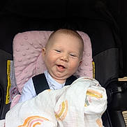 Elodie is registered to the contest to win money with this photo: baby, blanket, car_seat, child, smiling, infant, pink, white_clothing, fabric, happy, seat_belt, headrest, cute, face, person, toddler, comfort, indoors, safety, portrait