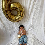 Jose is registered to the contest to win money with this photo: baby, child, cupcake, birthday_balloon, gold_balloon, number_6, white_backdrop, jeans, barefoot, smiling, frosting, messy, celebration, sitting, happy, portrait, indoor, playful, fun, cute