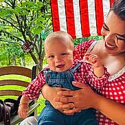 Nicolas joined the competition — help win amazing prizes! baby, child, parent, mother, smiling, happy, outdoor, portrait, family, infant, denim_overalls, checkered_shirt, red_clothing, flag, american_flag, greenery, trees, chair, seated, baby_carrier