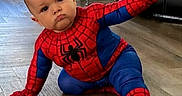 Nicolas is registered to the contest to win money with this photo: baby, child, spiderman, superhero, costume, red, blue, sitting, floor, indoors, portrait, face, serious_expression, outstretched_arm, playful, costume_design, wood_floor, chair, furniture, photography