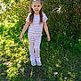 child, girl, outdoor, grass, sunlight, pajamas, striped_clothing, sneakers, greenery, bushes, daylight, nature, standing, casual, young, person, smile, hair_tied, playful, curious