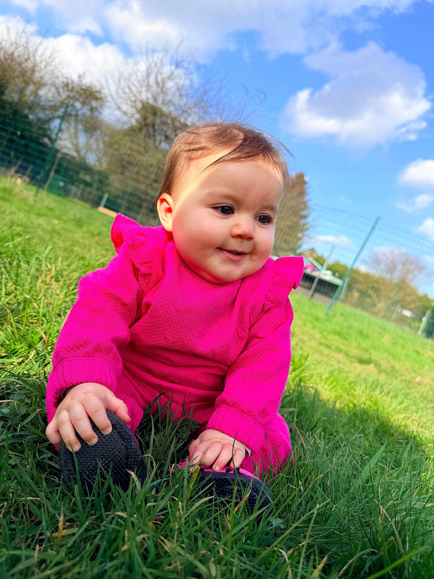 Atena a rejoint le concours — aidez-le/la à gagner de superbes lots ! baby, chubby_cheeks, clouds, cute, daylight, face, fence, grass, greenery, hand, infant, outdoors, park, pink_clothing, playful, portrait, shoe, sitting, sky, smile