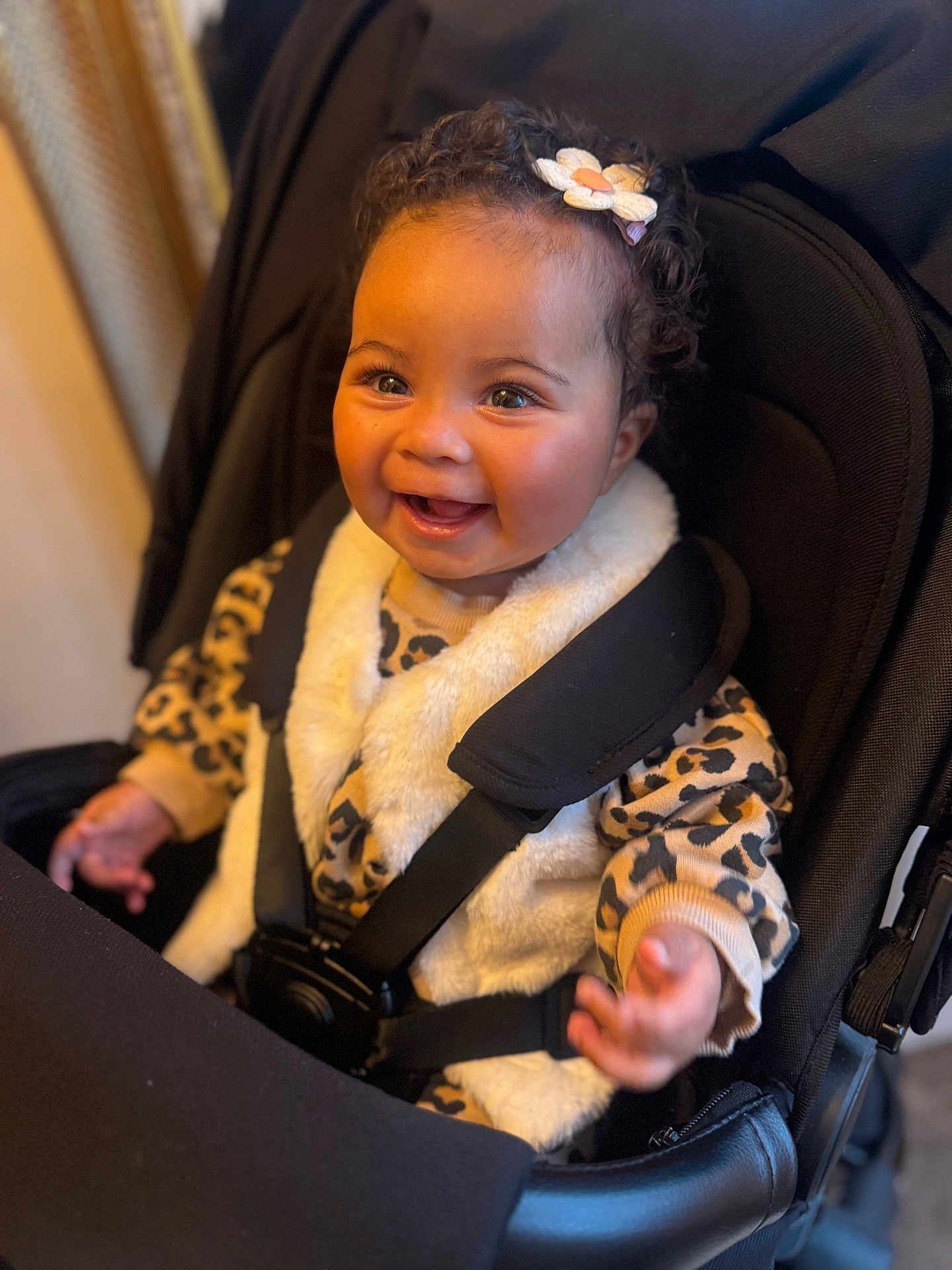 Myla is registered to the contest to win money with this photo: baby, child, smiling, stroller, leopard_print, fur_vest, curly_hair, hair_clip, happy, cute, infant, clothing, seatbelt, hands, portrait, indoor, person, face, expression, seat