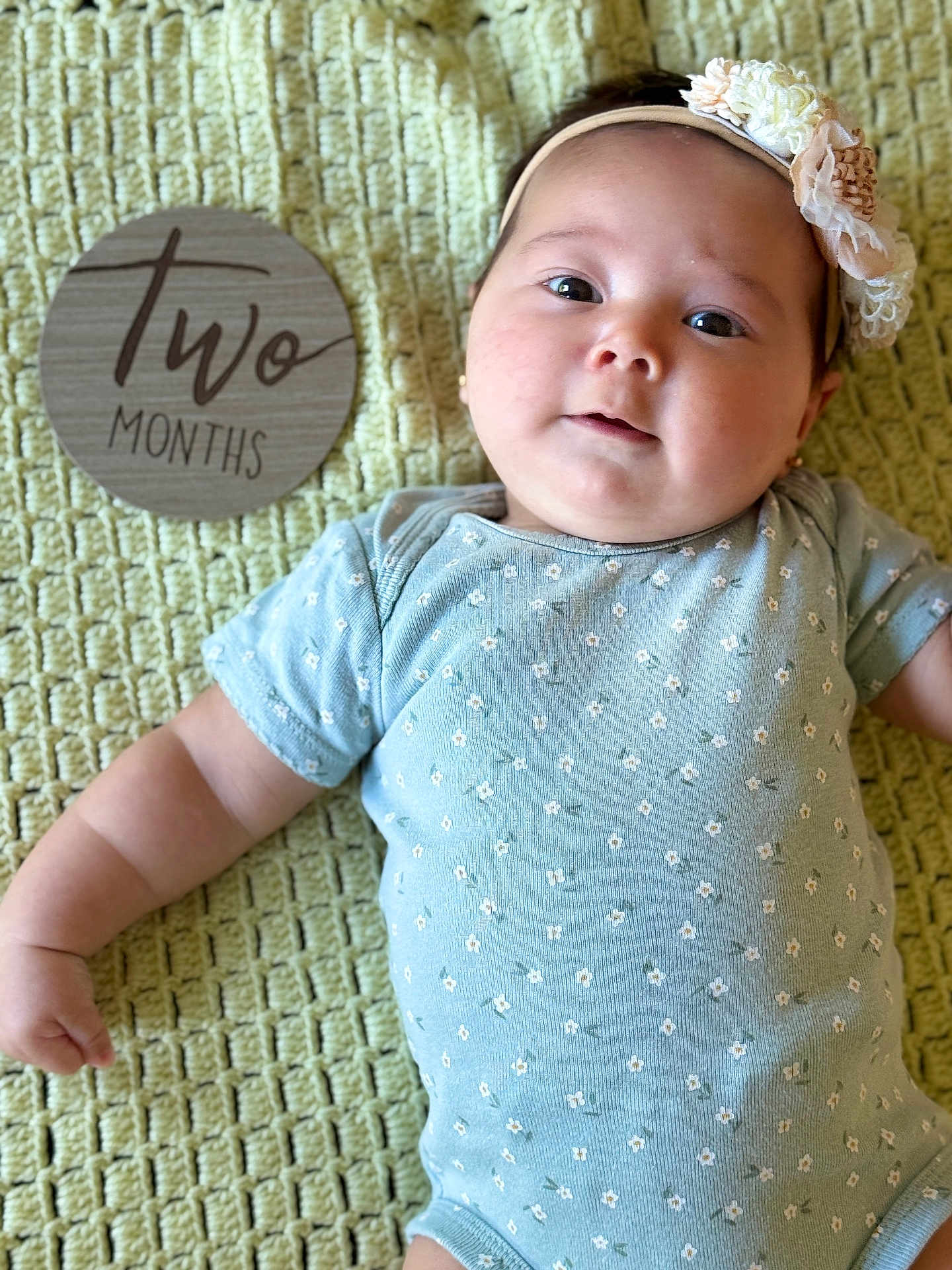 Gala is registered to the contest to win money with this photo: baby, infant, child, headband, flower, onesie, blanket, texture, sign, wood, milestone, portrait, cute, lying_down, skin, face, smile, small_person, happy, closeup