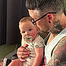 Bear is registered to the contest to win money with this photo: baby, infant, child, father, parent, portrait, pacifier, cuddling, holding, sitting, indoors, family, tattoos, glasses, beard, onesie, dinosaur_print, baby_clothing, eye_contact, photography
