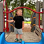 black_shirt, blue_slide, child, daytime, gray_shorts, green_sandals, happy, leisure, outdoor, park, person, play, playground, red_railing, slide, smiling, summer, toddler, trees, wooden_platform
