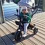 toddler, child, tricycle, beanie, jacket, outdoor, bridge, river, sunlight, shadow, vehicle, play, seat, handlebar, canopy, urban, nature, curious, casual_clothing, daytime
