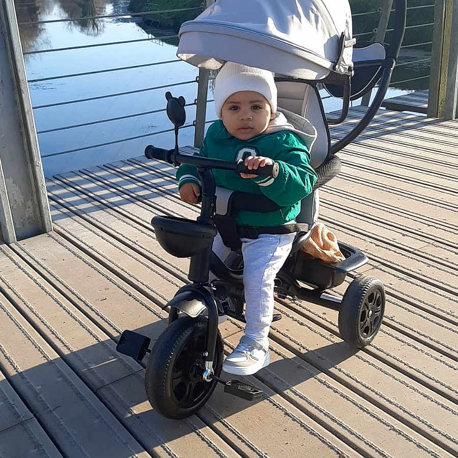 Khalil participe au concours pour gagner de l'argent avec cette photo : beanie, bridge, canopy, casual_clothing, child, curious, daytime, handlebar, jacket, nature, outdoor, play, river, seat, shadow, sunlight, toddler, tricycle, urban, vehicle