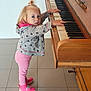 toddler, child, piano, musical_instrument, hoodie, pink_leggings, pink_sneakers, hair_tie, standing, curious, indoors, tile_floor, wooden_piano, keys, cute, portrait, small_hands, playing, young_child, casual_clothing