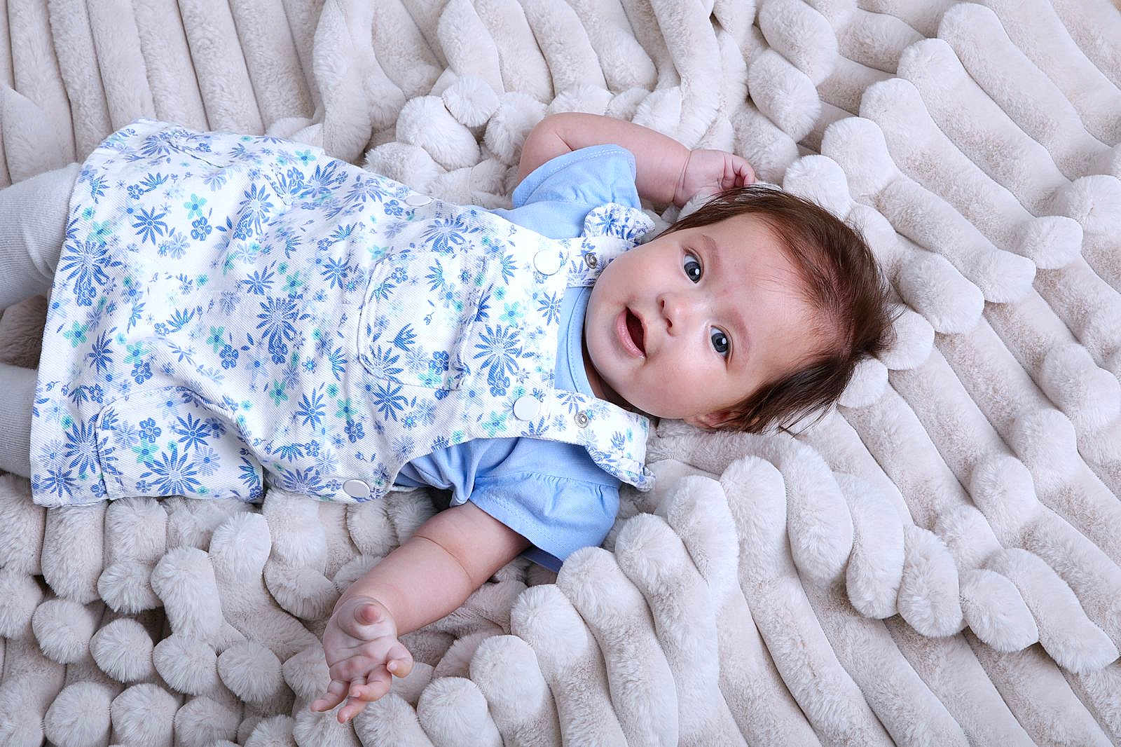 Deyna is registered to the contest to win money with this photo: baby, infant, child, blue_clothing, floral_pattern, blanket, plush, soft_texture, curious_expression, lying_down, cute, portrait, indoors, skin, hand, face, head, young, smiling, toddler