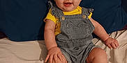 Hope is registered to the contest to win money with this photo: baby, child, smile, bow, headband, denim_overalls, yellow_shirt, socks, paws, bed, pillow, blue_pillow, happy, cute, infant, person, indoor, clothing, sitting, portrait