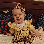 Hope is registered to the contest to win money with this photo: baby, child, smiling, bed, pillow, yellow_clothing, headband, leopard_print, sitting, happy, indoor, cute, infant, person, text_on_clothing, footwear, blanket, home, portrait, apparel
