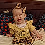 baby, child, smiling, bed, pillow, yellow_clothing, headband, leopard_print, sitting, happy, indoor, cute, infant, person, text_on_clothing, footwear, blanket, home, portrait, apparel