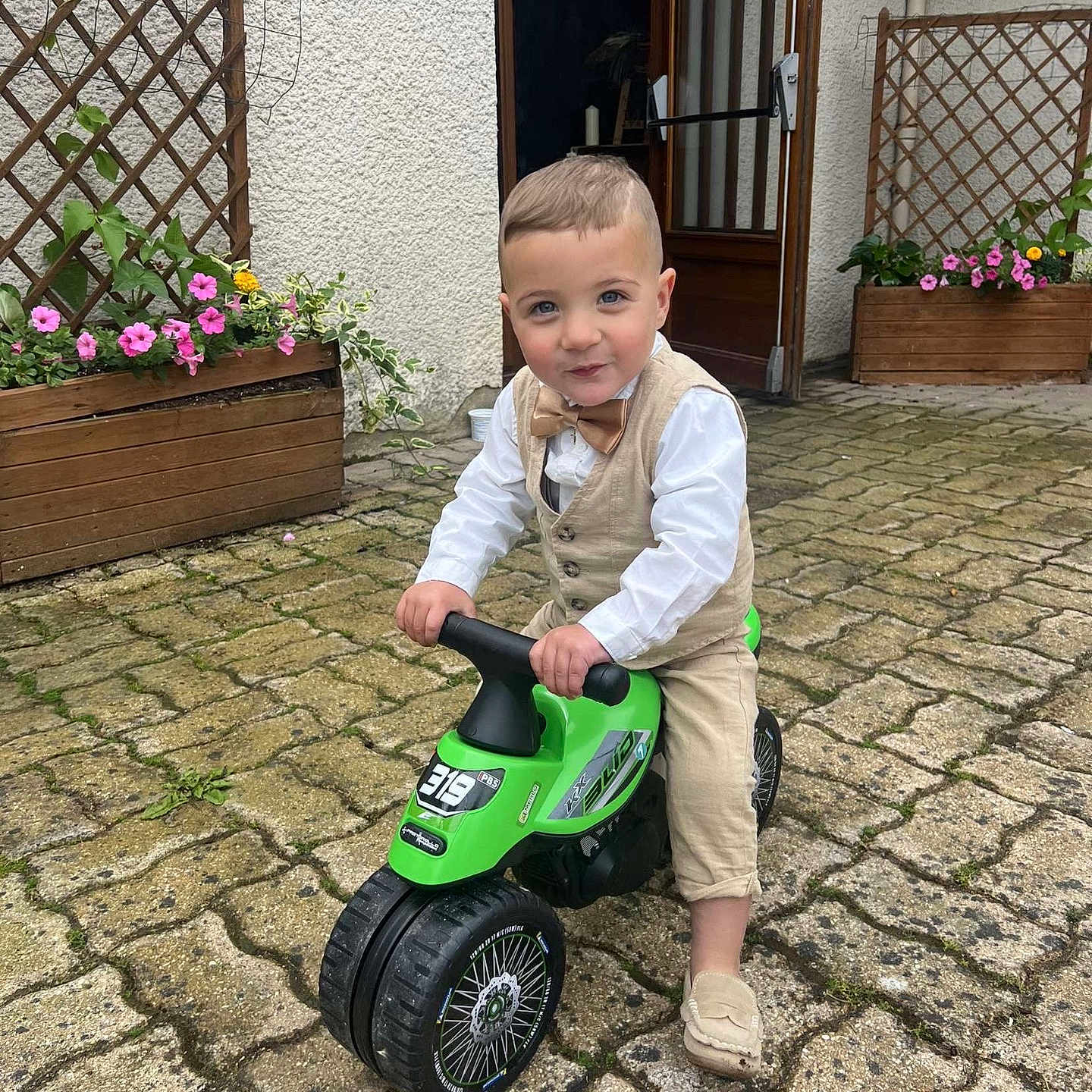 Maël participe au concours pour gagner de l'argent avec cette photo : bow_tie, child, clothing, daylight, doorway, flowers, garden, green, outdoor, paved_ground, person, plant, play, shoes, smile, toddler, toy_motorcycle, vest, wooden_planter, young_child