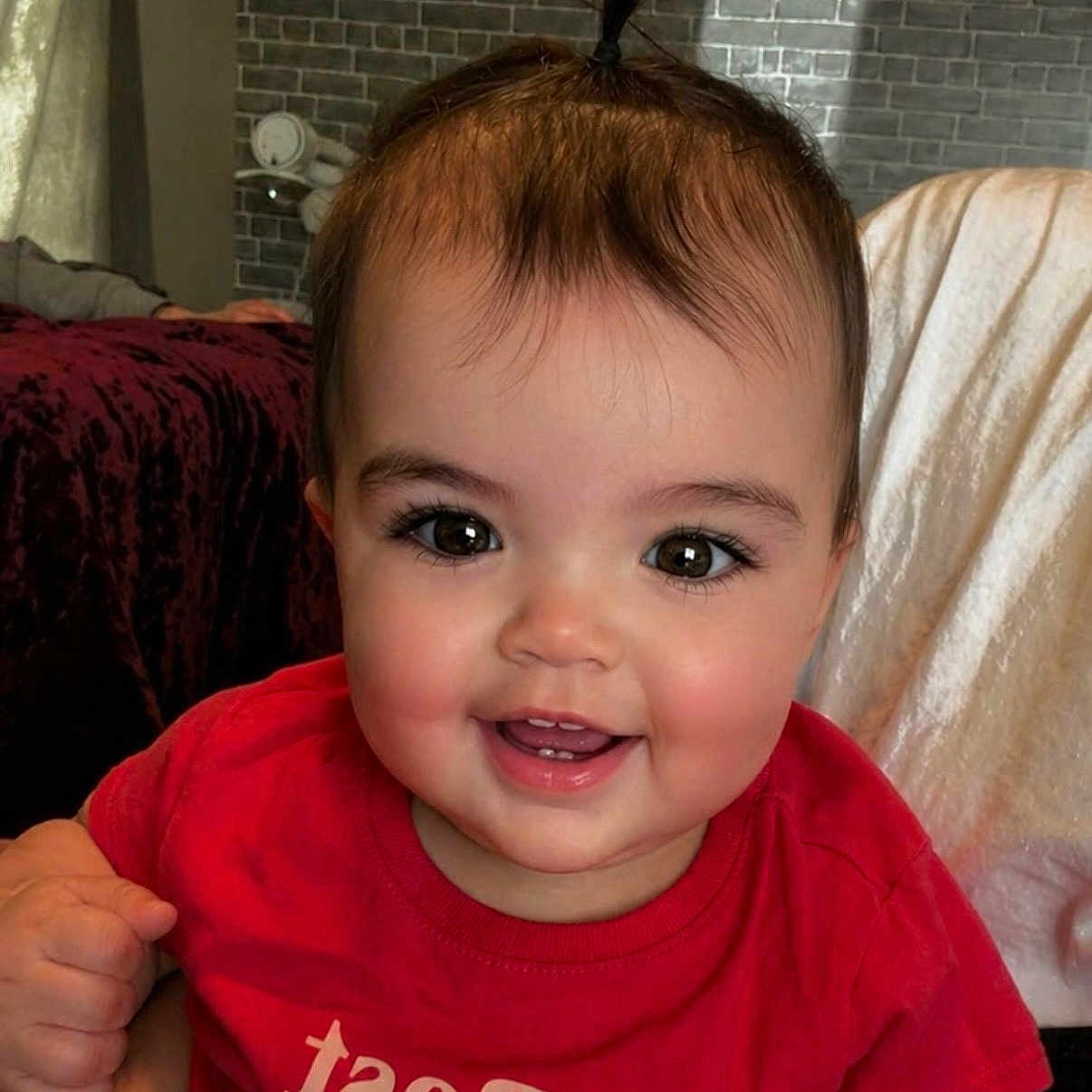 Kinsley joined the competition — help win amazing prizes! baby, child, smiling, red_shirt, ponytail, face, indoors, brick_wall, furniture, happy, cute, person, portrait, toddler, clothing, skin, head, eyes, mouth, hand