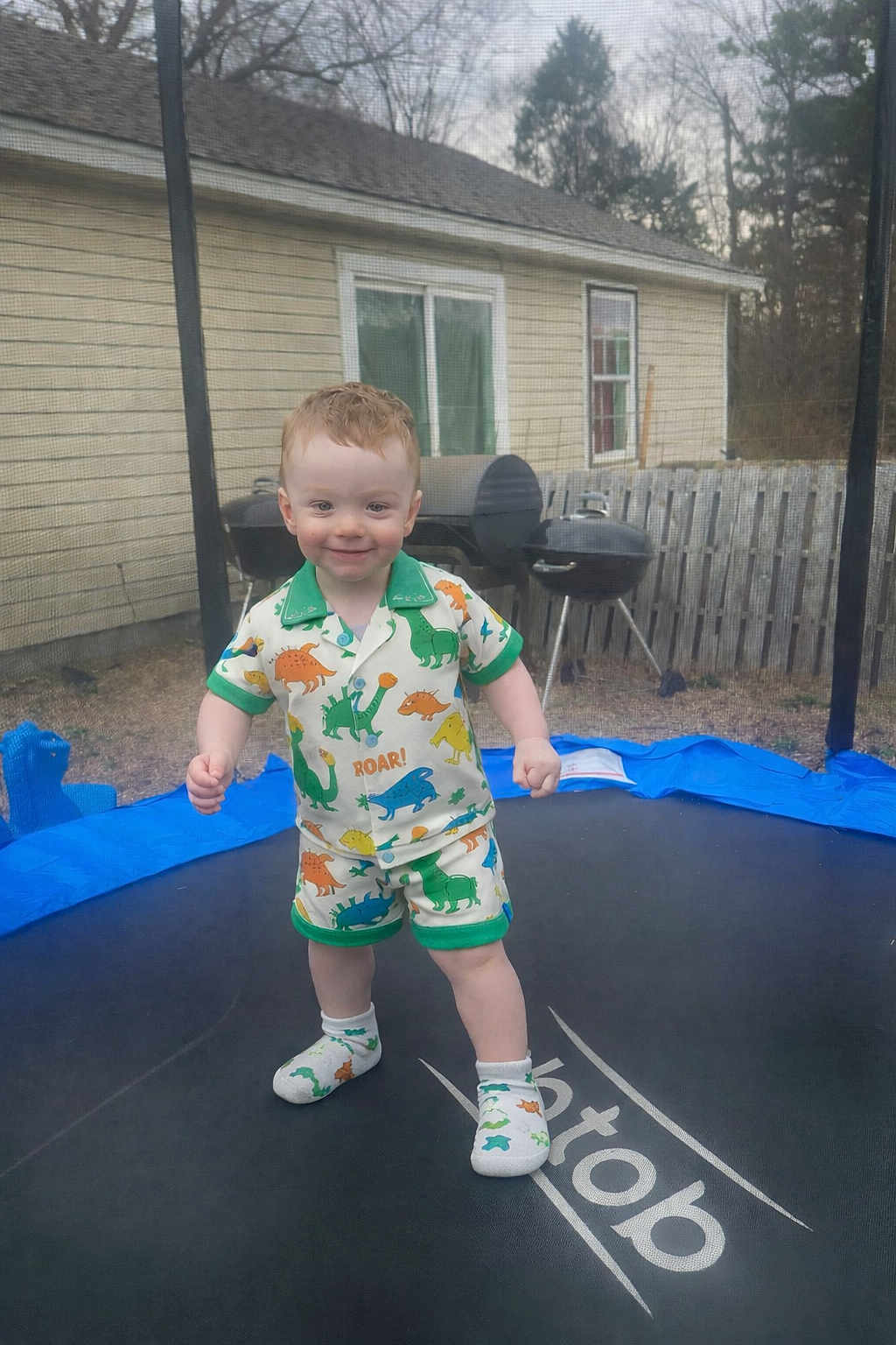Landyn Scott is registered to the contest to win money with this photo: child, toddler, trampoline, dinosaur_pajamas, socks, standing, smiling, backyard, house, grill, fence, safety_net, grass, outdoor, portrait, red_hair, play, young_child, happy, clothing