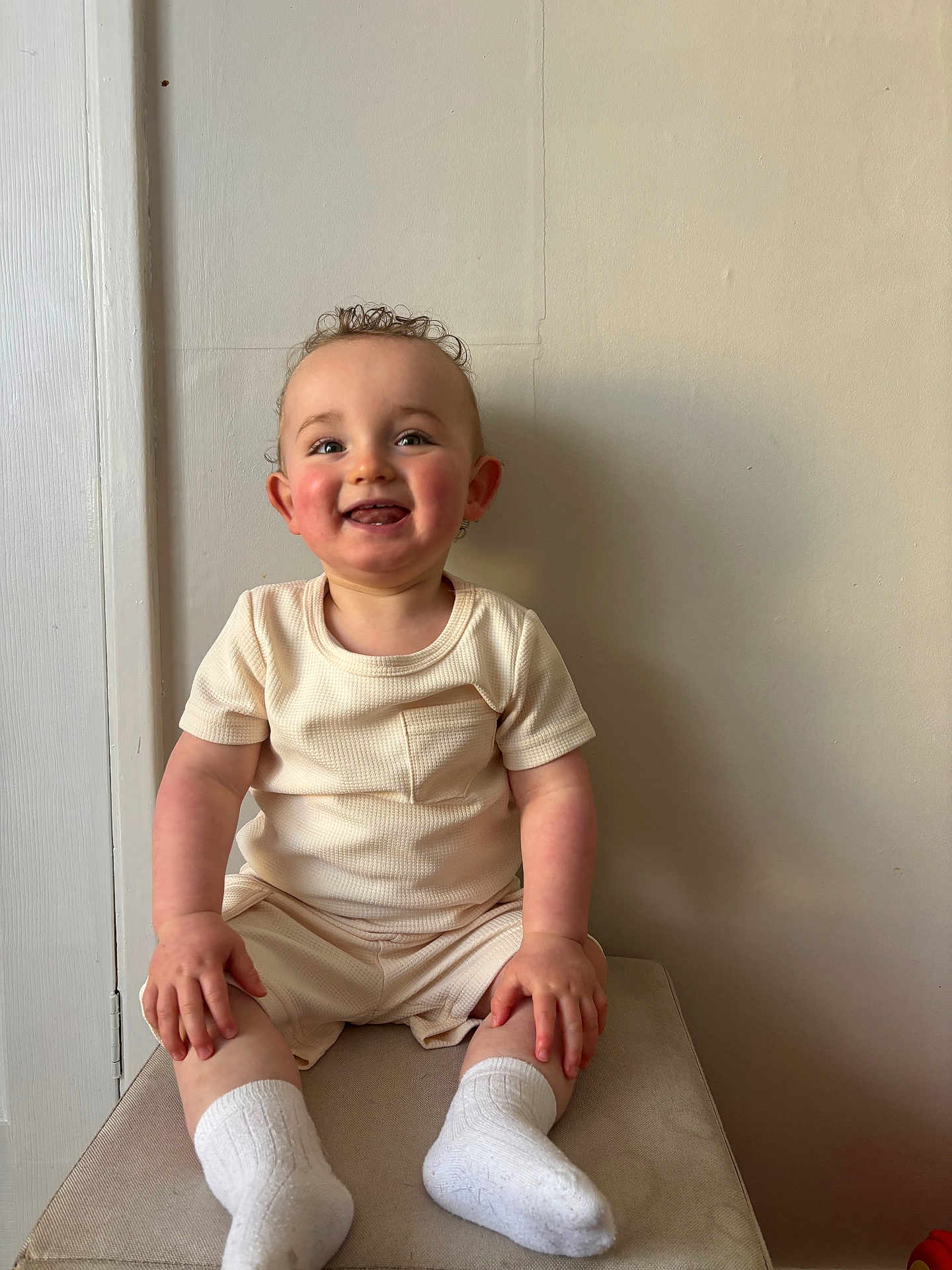 Rowan is registered to the contest to win money with this photo: baby, child, smiling, happy, curly_hair, sitting, white_socks, cream_clothing, indoor, cushion, face, person, cute, portrait, young_child, wall, furniture, hand, feet, skin