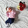 adorable, baby, cozy, curious_look, cute, decorative_wrapping, flower_bouquet, headband, heart_pattern, indoor, infant, lying_down, navy_pants, pink_shirt, portrait, red_flowers, small_hands, soft_lighting, white_blanket, white_flowers