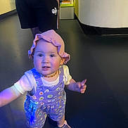 Amélia a rejoint le concours — aidez-le/la à gagner de superbes lots ! background, casual_clothing, child, colorful, curious, floor, footwear, hat, indoor, infant, overalls, people, person, pink, portrait, reaching, shorts, standing, toddler, young_child