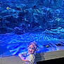 Amélia participe au concours pour gagner de l'argent avec cette photo : aquarium, baby, blue_light, child, curious, cute, display, exhibit, fish, indoor, information_plaques, person, purple_bonnet, purple_outfit, rocks, smile, standing, toddler, underwater, water