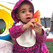 Natasha is registered to the contest to win money with this photo: baby, bib, child, curious, daylight, face, grass, hands, hood, open_mouth, orange_toy, outdoor, patterned_clothing, pink_clothing, play_structure, playground, portrait, sitting, toddler, toy