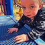 Levi is registered to the contest to win money with this photo: toddler, child, playground, outdoor, blue, metal, climbing, hands, face, eyes, jacket, sunlight, trees, play_equipment, curious, young_child, nature, daylight, park, exploring