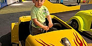 Levi is registered to the contest to win money with this photo: child, toy_car, yellow, red_flames, indoor, play_area, clothing, short_hair, person, floor, car, activity_center, playground, young, curious_expression, seat, hand, wall, sign, table