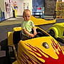 child, toy_car, yellow, red_flames, indoor, play_area, clothing, short_hair, person, floor, car, activity_center, playground, young, curious_expression, seat, hand, wall, sign, table