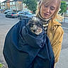 woman, dog, jacket, street, suburban, car, house, sidewalk, outdoor, pet, animal, portrait, holding, clothing, curly_hair, blond_hair, tree, casual, daytime, cute