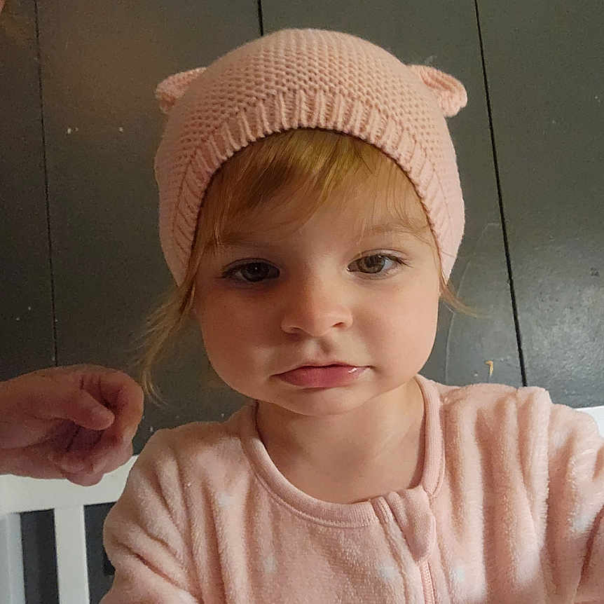 Tiffany is registered to the contest to win money with this photo: baby, child, closeup, clothing, curious, cute, expression, face, hand, headwear, indoor, knit_hat, looking, person, pink_hat, portrait, railing, soft_lighting, toddler, young_child