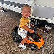 Taysem participe au concours pour gagner de l'argent avec cette photo : toddler, child, toy_car, orange, black, yellow_shirt, striped_overalls, white_shoes, gravel, trailer, wheel, outdoor, smiling, cute, person, small_shoes, play, vehicle, ground, daylight