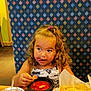 basket, booth, bowl, child, chips, curly_hair, cute, dining, drink, food, girl, hair_tie, patterned_upholstery, plastic_cup, playful, restaurant, salsa, snack, straw, table