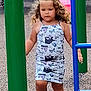 blue_pole, casual_clothing, child, concrete_ground, curly_hair, daylight, dress, focused_expression, green_pole, outdoor, patterned_clothing, person, pink_slide, play_equipment, playground, playtime, slide, summer, walking, young_child