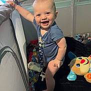 Archer is registered to the contest to win money with this photo: baby, playpen, smile, standing, toy, blanket, indoors, child, cute, happy, blue_clothing, stuffed_animal, face, person, infant, light, shadow, hand, foot, nursery