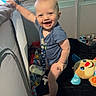 baby, playpen, smile, standing, toy, blanket, indoors, child, cute, happy, blue_clothing, stuffed_animal, face, person, infant, light, shadow, hand, foot, nursery
