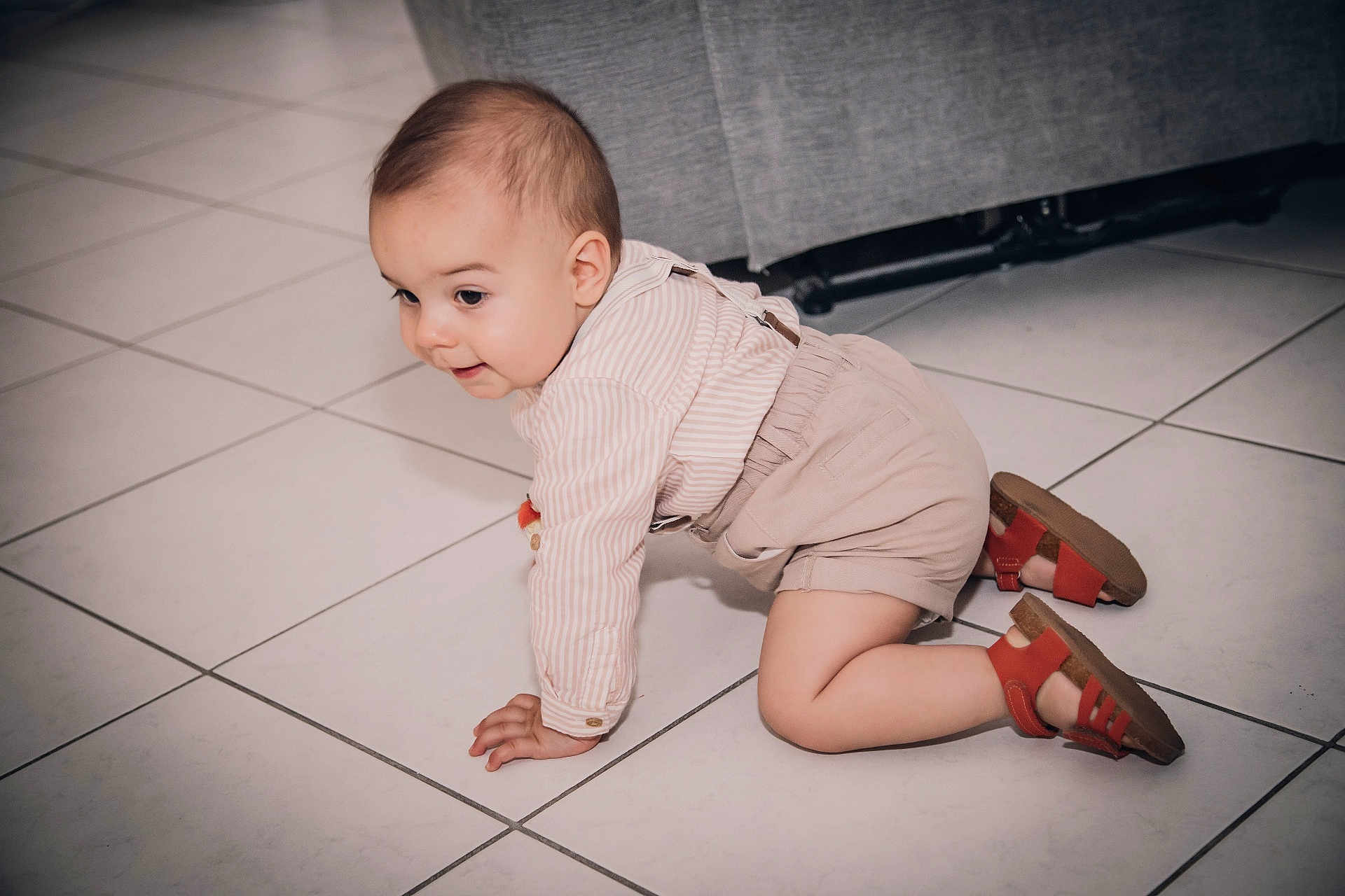 Lëlyo a rejoint le concours — aidez-le/la à gagner de superbes lots ! baby, beige_shorts, child, clothing, couch, crawling, curious, face, floor, footwear, hands, head, home_interior, indoors, knees, long_sleeve_shirt, person, red_sandals, tile_floor, young_child