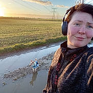 Louna participe au concours pour gagner de l'argent avec cette photo : person, dog, headphones, outdoor, field, sunset, puddle, muddy_path, jacket, small_dog, power_lines, sky, clouds, grass, reflection, walking, nature, casual_clothing, selfie, blue_jacket