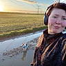 person, dog, headphones, outdoor, field, sunset, puddle, muddy_path, jacket, small_dog, power_lines, sky, clouds, grass, reflection, walking, nature, casual_clothing, selfie, blue_jacket