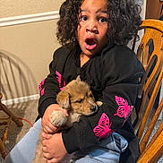 Bear joined the competition — help win amazing prizes! black_sweatshirt, blue_pants, carpet, casual_clothing, chair, child, curly_hair, cute, dog, holding, home, indoor, light_skin, pet, pink_butterfly_print, portrait, puppy, sitting, surprised_expression, wooden_furniture
