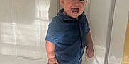 Jack is registered to the contest to win money with this photo: toddler, child, smiling, blue_clothing, shorts, shirt, white_sneakers, standing, indoor, door, carpet, shadow, happy, portrait, cute, young_child, person, casual_wear, light, expression
