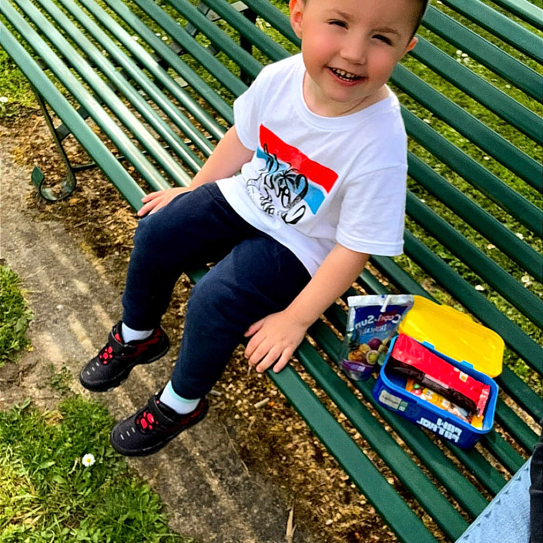 Tiago participe au concours pour gagner de l'argent avec cette photo : bench, boy, cap, casual, child, daylight, footwear, grass, green, happy, jeans, nature, outdoor, park, person, shoes, sitting, smiling, snacks, tshirt