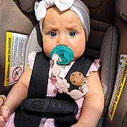 Gracia is registered to the contest to win money with this photo: baby, infant, car_seat, pacifier, blue_eyes, white_bow, pink_clothing, safety_harness, child, cute, portrait, seatbelt, baby_accessory, closeup, adorable, young_child, car_safety, straps, indoor, expression