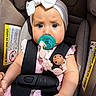 baby, infant, car_seat, pacifier, blue_eyes, white_bow, pink_clothing, safety_harness, child, cute, portrait, seatbelt, baby_accessory, closeup, adorable, young_child, car_safety, straps, indoor, expression