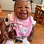Khyla is registered to the contest to win money with this photo: baby, child, smiling, happy, indoor, bib, pacifier, hair_buns, person, wooden_floor, chair, table, clothing, hand, face, cute, infant, holding, seated, room