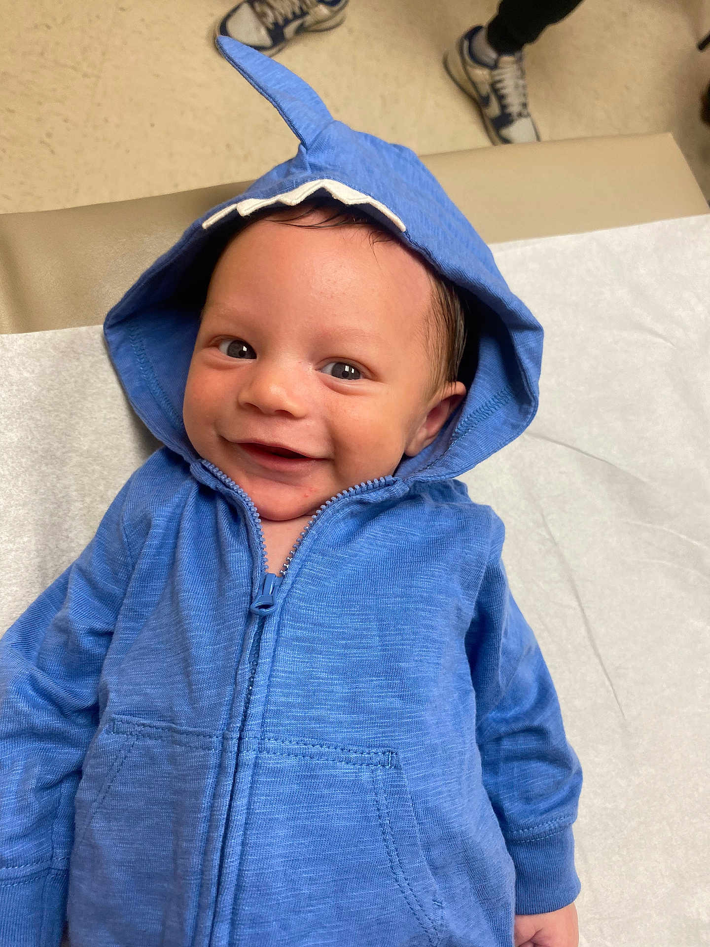 Jahkarri is registered to the contest to win money with this photo: baby, blue_hoodie, smiling, infant, face, hood, medical_table, sneakers, person, young_child, cute, happy, clothing, indoor, portrait, child, casual_wear, expression, healthcare, closeup