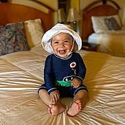 Ezekiel is registered to the contest to win money with this photo: baby, smiling, hat, white_hat, swimsuit, navy_blue, crocodile_design, bed, bedroom, pillow, headboard, furniture, happy, sitting, infant, child, indoors, foot, cute, portrait