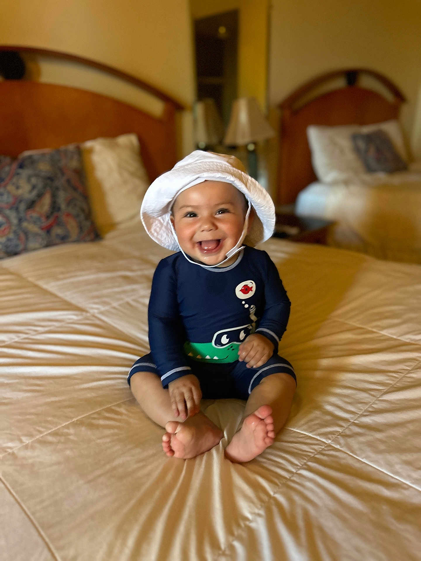 Ezekiel is registered to the contest to win money with this photo: baby, smiling, hat, white_hat, swimsuit, navy_blue, crocodile_design, bed, bedroom, pillow, headboard, furniture, happy, sitting, infant, child, indoors, foot, cute, portrait