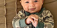 Luke is registered to the contest to win money with this photo: baby, infant, smiling, camouflage, onesie, clothing, hands, face, eyes, bed, quilt, fabric, cute, portrait, child, lying_down, skin, head, happy, young