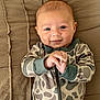 baby, infant, smiling, camouflage, onesie, clothing, hands, face, eyes, bed, quilt, fabric, cute, portrait, child, lying_down, skin, head, happy, young